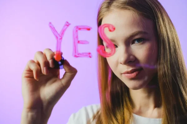 Teenage girl writing the word 'Yes' on glass with marker in studio setting.