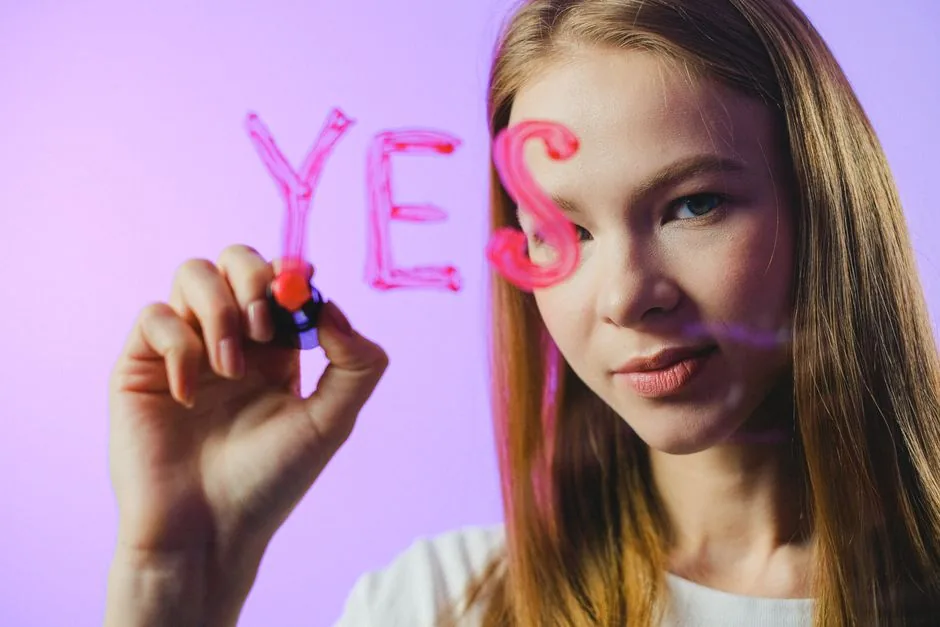 Teenage girl writing the word 'Yes' on glass with marker in studio setting.