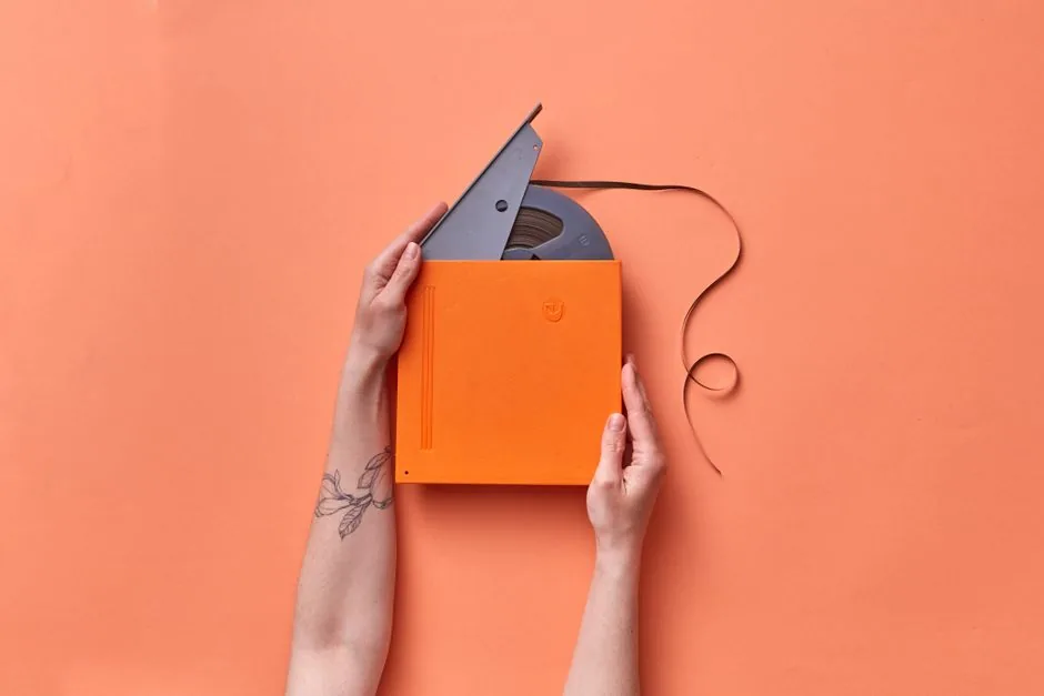 A pair of hands holding a vintage film tape in an orange box against a matching orange background.